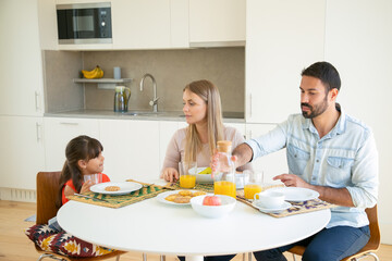 Parents couple and girl having breakfast, sitting at dining table with fruit, biscuits and orange juice, talking and eating. Medium shot. Family morning concept