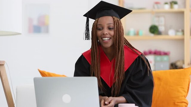 African American female student is using laptop while sitting on sofa in apartment room spbi. Beautiful lady wearing cap and gown looking at computer screen with smile during video conference on couch