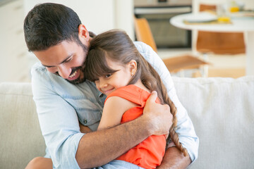 Joyful dad sitting with his little girl on couch, hugging and cuddling her. Happy father enjoying...