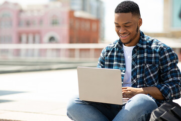 Glad black man using laptop, sitting in the city downtown