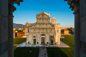 Lovely photo of the Pisa Cathedral façade with the Leaning Tower in the background, taken from a window of the Pisa Baptistery, flanked by two columns. Tourists enjoy the Piazza dei Miracoli by dusk.