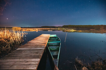 Belarus, Eastern Europe. Real Night Sky Stars Above Old Pier With Moored Wooden Fishing Boat. Natural Starry Sky And Countryside Landscape With Lake River In Early Spring Night. Russian Nature