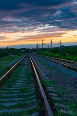 Fototapeta na wymiar Railroad against the backdrop of sky and sunset clouds with green grass in the foreground. Beautiful sunset with orange sunbeams on a background of blue clouds. Leaving road concept.