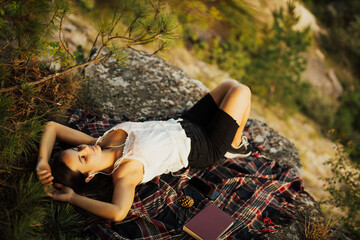 Young stylish hipster woman lying on the rock on the mountain. Smiling model enjoy her weekends. Female listening to music via headphones.Top view.