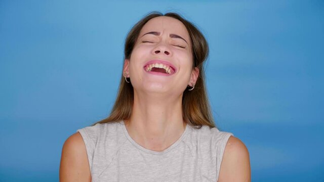Beautiful cheerful woman in gray T-shirt laughs, looks around on blue background with copy space. 4K slow motion footage