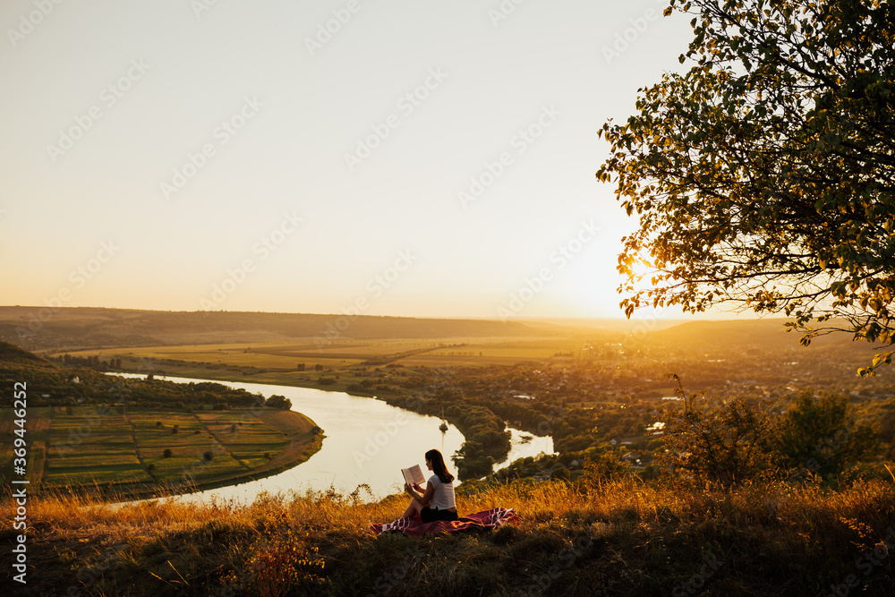 Wall mural Young woman tourist sitting on hill with river on background and reading a book at sunset in the mountains. She reading carefully staring at the open book. Beautiful landscape.