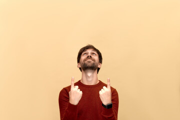 Look up. Photo of young man with beard wearing sweater, points at copy space from above on yellow wall as shows something pleasant, has smiling look, advertises product.