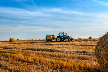 A tractor uses a trailed bale machine to collect straw in the field and make round large bales