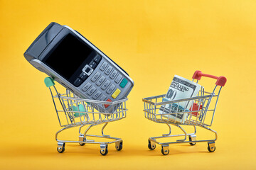 Mini shopping cart with bank cards inside, dollar banknotes and terminal on yellow background. Trolley and money. Financial crisis or shopaholic concept. Flat lay, copy space.