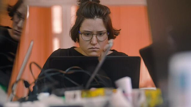 sad calm woman in glasses sitting at office working by the laptop irrl. making grimace. company worker at messy workplace. female manager working with computer data. tired
