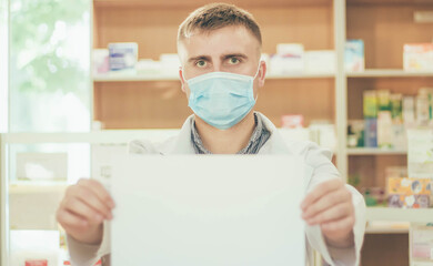 Fototapeta na wymiar A man pharmacist in a medical mask and a white coat against the background of a pharmacy counter holds a white piece of paper in front of him