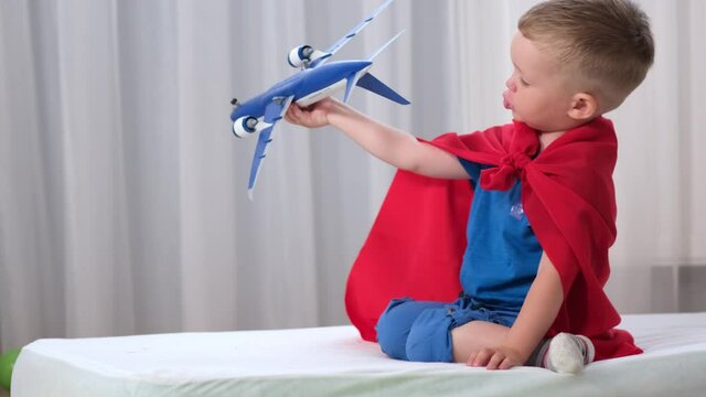 Blond caucasian boy super hero kid is playing with toy air plane in children's room dressed in red raincoat and blue t-shirt. Boy dreams of becoming an air pilot of an airplane.