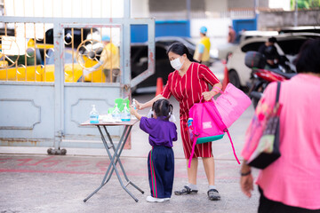 Mother is taking her daughter to wash her hands with alcohol gel after returning from school. Mother holds a mattress and pink bag. Families wear masks to protect against viruses and poisonous dust.
