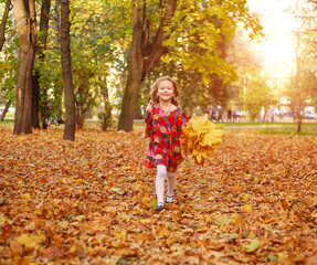 Portrait of happy joyful running child girl on fallen yellow autumn foliage in autumn city forest park in rays of sunset light