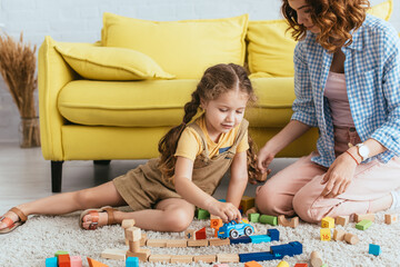 young nanny sitting near adorable kid playing with toy car near multicolored blocks on floor