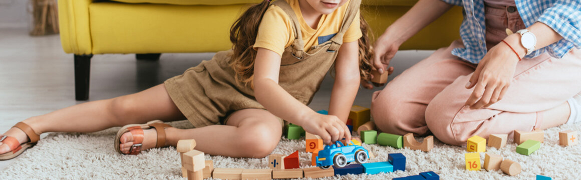 cropped view of babysitter near child playing with toy car near multicolored blocks on floor, horizontal image