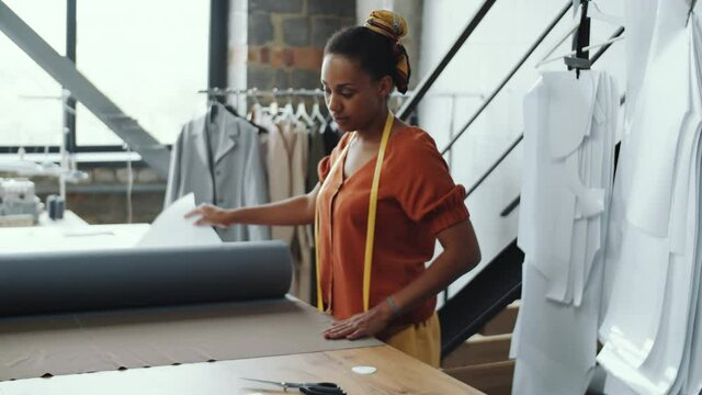 Young African American seamstress unrolling fabric on worktable, applying paper sewing pattern and outlining it with chalk while working in atelier