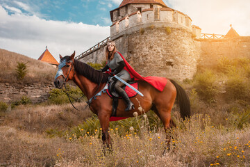 A beautiful girl with a sword in a medieval fantasy suit. A woman in a chain mail, crown, on a horse in combat ammunition. A girl riding a horse against the backdrop of the fortress. © Valeriia