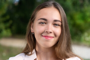 Fototapeta premium Portrait of a young girl outdoors on a summer evening. Sincere smile. Beautiful smiling teenage girl in a blue blouse, against a green summer park.