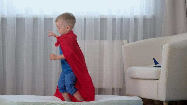 Slow motion blond caucasian boy super hero kid in red raincoat of hero having fun and jumping in room on mattress as on trampoline up, boy having fun and enjoying his childhood, travel concept