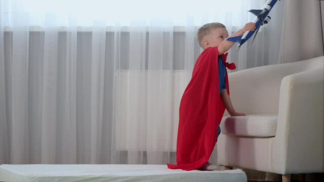 Blond caucasian boy super hero kid playing with toy air plane, jumping on mattress, trampoline with toy in his hands dressed in red raincoat of hero and blue t-shirt in children's room.