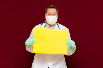 Young handsome doctor holding yellow blank banner. Place your text advertisement.burgundy wall.