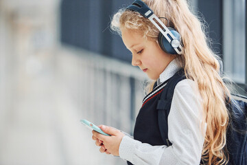 Cute little school girl in uniform with headphones and phone indoors