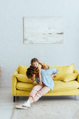 young nanny piggybacking happy kid while sitting on yellow sofa