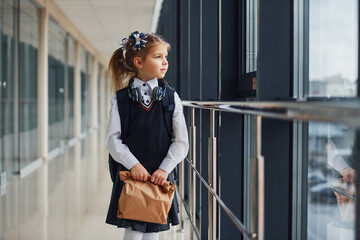 Fototapeta premium Young little school girl in uniform standing in hallway with package of dinner in hands