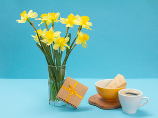Bouquet of yellow daffodils in vase with gift box and a cup of coffee on a blue background.