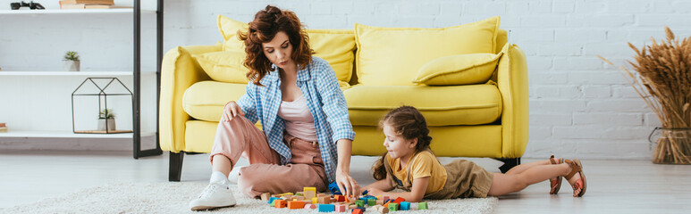 panoramic concept of young babysitter and adorable kid playing with multicolored blocks on floor in living room