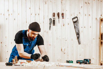 A dark-haired man with a beard and in overalls treating a wooden bar with a black jack plane,  in the background a lot of wooden boards. Work with wooden .