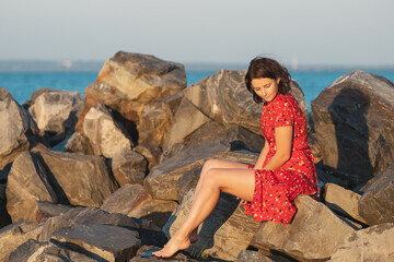Young beautiful woman in a light casual red dress  resting and posing on rocks on a sandy beach near a blue sky on a summer day, beautiful sea against a blue sky