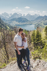 Fototapeta na wymiar Lovely caucasian couple in white T-shirts stand on the top & embrace on background of Alpsee lake, green forest, rock mountains with snow peaks & blue sky. Bavaria. Germany