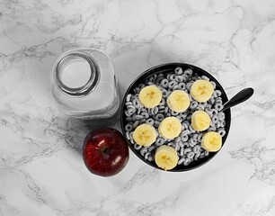 Looking down at bowl of cereal with banana slices, red apple and glass of milk. 