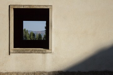A view of mountains on the horizon with a blue sky and green trees seen through a window within another window in a clean old wall of an abandoned building on the outskirts of the city.