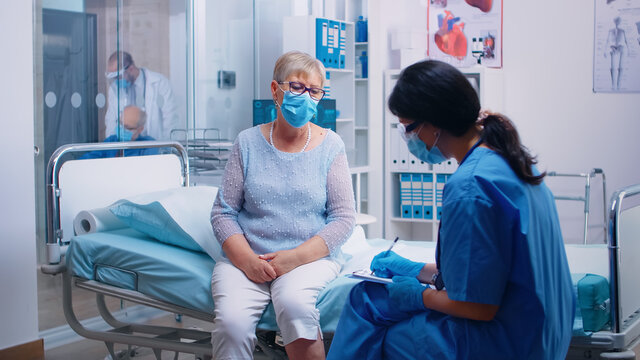 Nurse in protective equipment questioning senior lady patient who wears a mask. Modern private hospital, clinic or healthcare facility. Doctor appointment during coronavirus outbreak