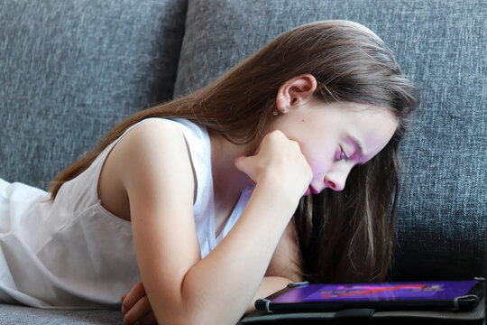 girl 9 years old looking in digital tablet. pink light reflects on her face. Girl lying on her stomach on the couch uses lapot at home.