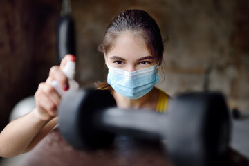 Girl cleaning of sport equipments of gym with  disinfecting spray. Woman making disinfection of dumbbells with sanitizer during epidemic. Protection and prevention from infection.