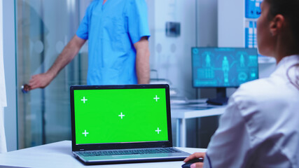 Chroma key on laptop in medical clinic used by doctor wearing white coat and nurse with blue uniform opening cabinet glass door. Medic wearing uniform using notebook with copy space on display in