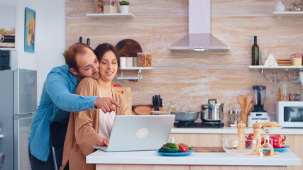 Wife reading on laptop in kitchen while husband is hugging her. Husband and wife cooking recipe food. Happy healthy together lifestyle. Family searching for online meal. Health fresh salad