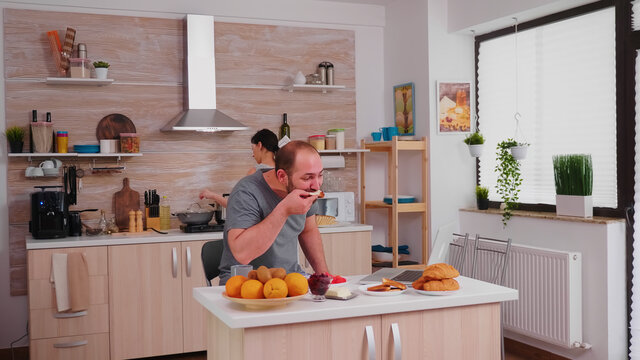 Freelancer drinking coffee and working on laptop during breakfast in kitchen. Entrepreneur in pajamas working on a laptop from home in kitchen.