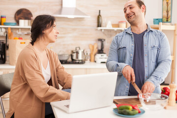 Couple preparing organic salad in kitchen. Wife using laptop. Man laughing. Man helping woman to prepare healthy organic dinner, cooking together. Romantic cheerful love relationship