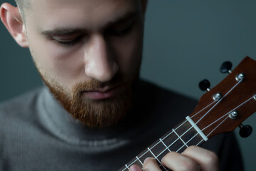 close-up of a young bearded guy playing the ukulele, on a gray background. Studio portrait of a...