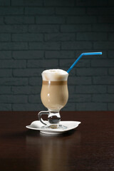Coffee cappuccino in a glass Cup with a tube on a wooden table against a brick wall. Photos for restaurant, cafe and bar menus
