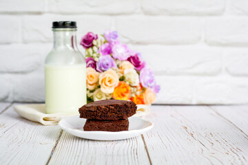 Obraz na płótnie Canvas Piece of brownie cake on white small plate with bottle of fresh milk on wooden table and with brick wall with copy space.