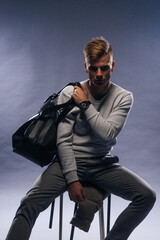Informally ( casual ) dressed blonde model wearing a white sweater and holding a black man bag is posing on a white background while sitting on a chair.