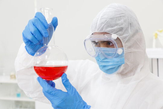 An asian man in a protective suit  (PPE Suit) and mask holds round bottom flask and vaccine. Vaccine from, flu, coronavirus, ebola, TB. Biological hazard. Epidemic of the Covid-19.