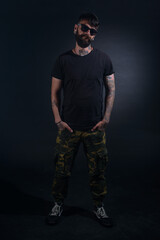 Bearded male model with tattoos and hands in his pocket, dressed in a black t-shirt and sunglasses poses over black background.