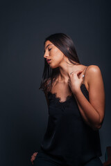 Fototapeta premium Sexy brunette wearing a black tank top and black pants posing while sitting on the top edge of a chair inside a studio on a dark background